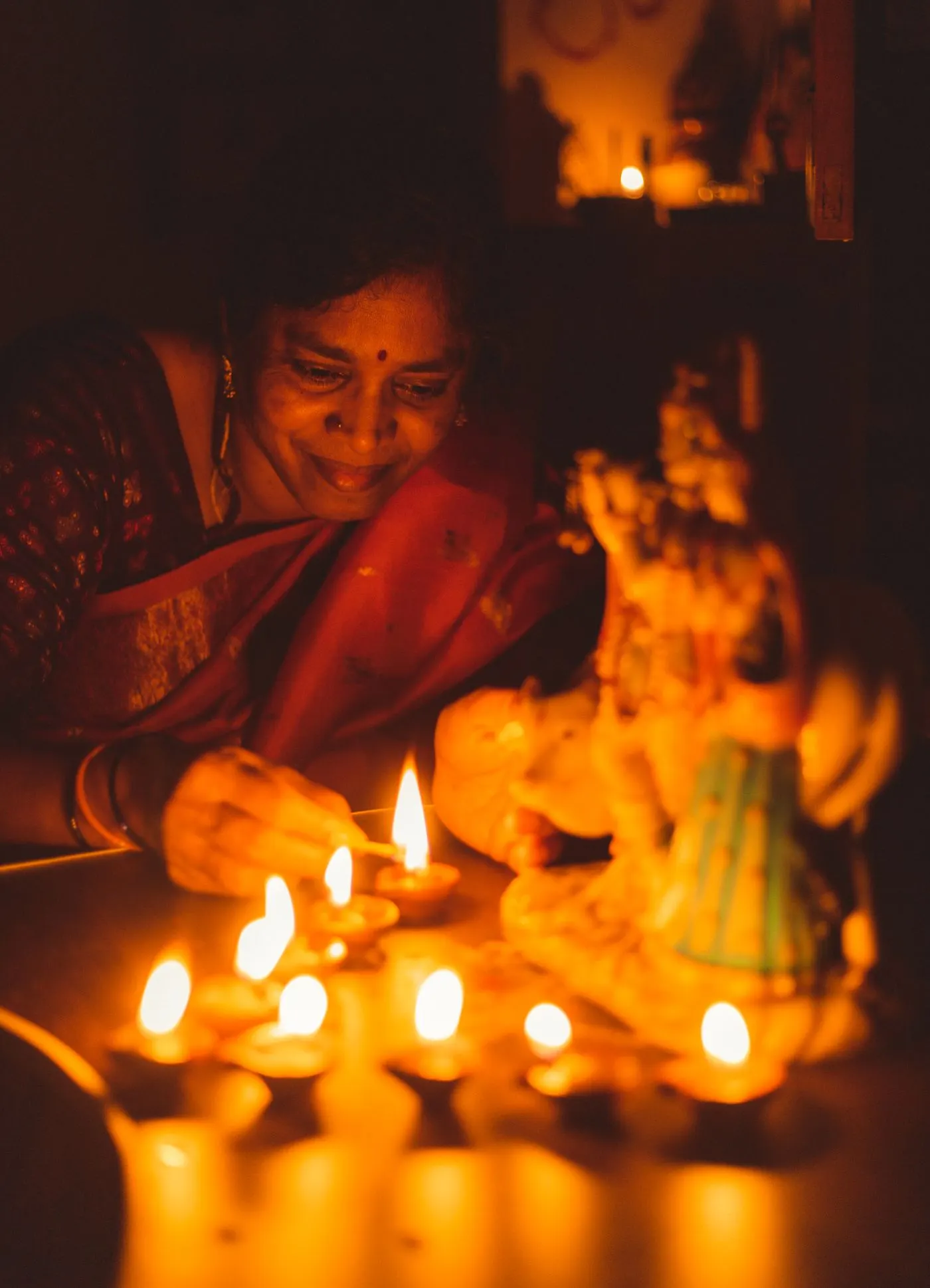 A woman in a red saree lighting clay diyas at home, with a small shrine beside her